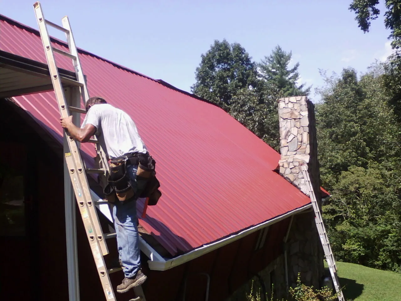 Roofer on a ladder beside a red metal roof and stone chimney — Soffit & Fascia Repair by Roofing Brothers in Hood River
